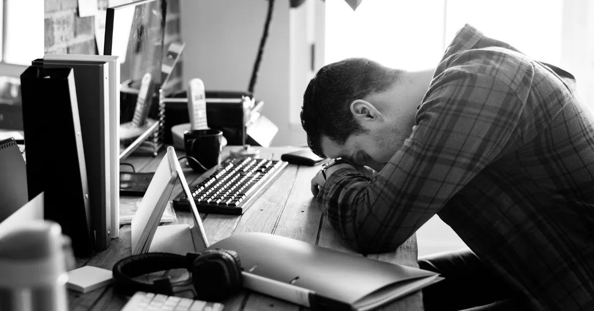 仕事上での精神的なプレッシャーに押しつぶされそうな男性。デスクに倒れ込みそうになっている