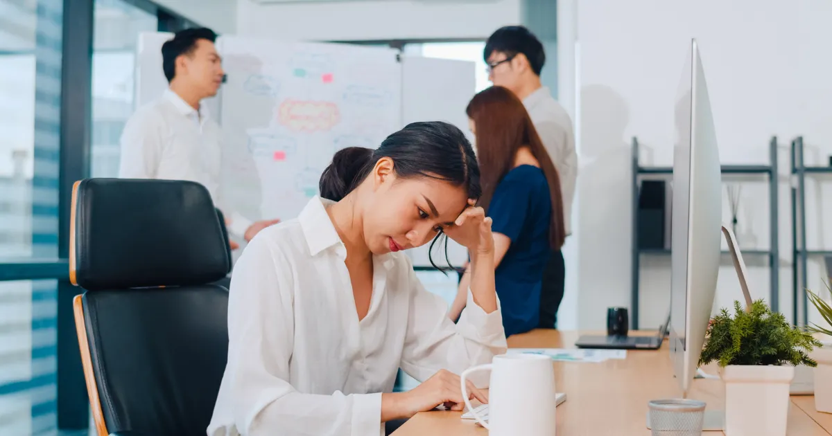 職場で同僚たちが談笑するなか、人間関係の悩みを抱え、一人デスクで頭を抱えて孤立している女性の様子